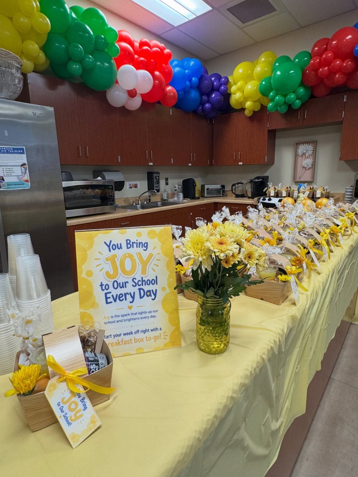 Yellow themed appreciation table with flowers, gift boxes, and a colorful balloon arch.