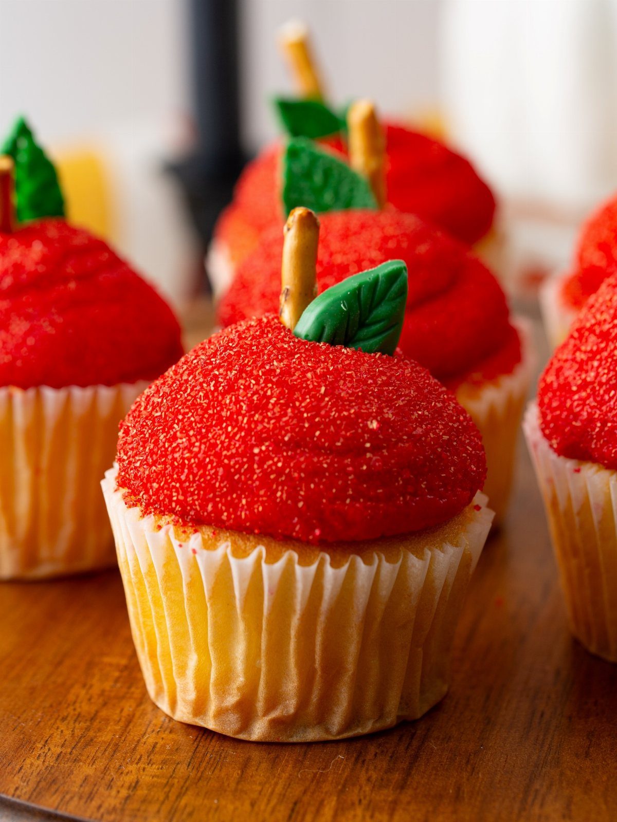 Apple cupcakes with red sugar frosting, pretzel stems, and green leaves on wood.