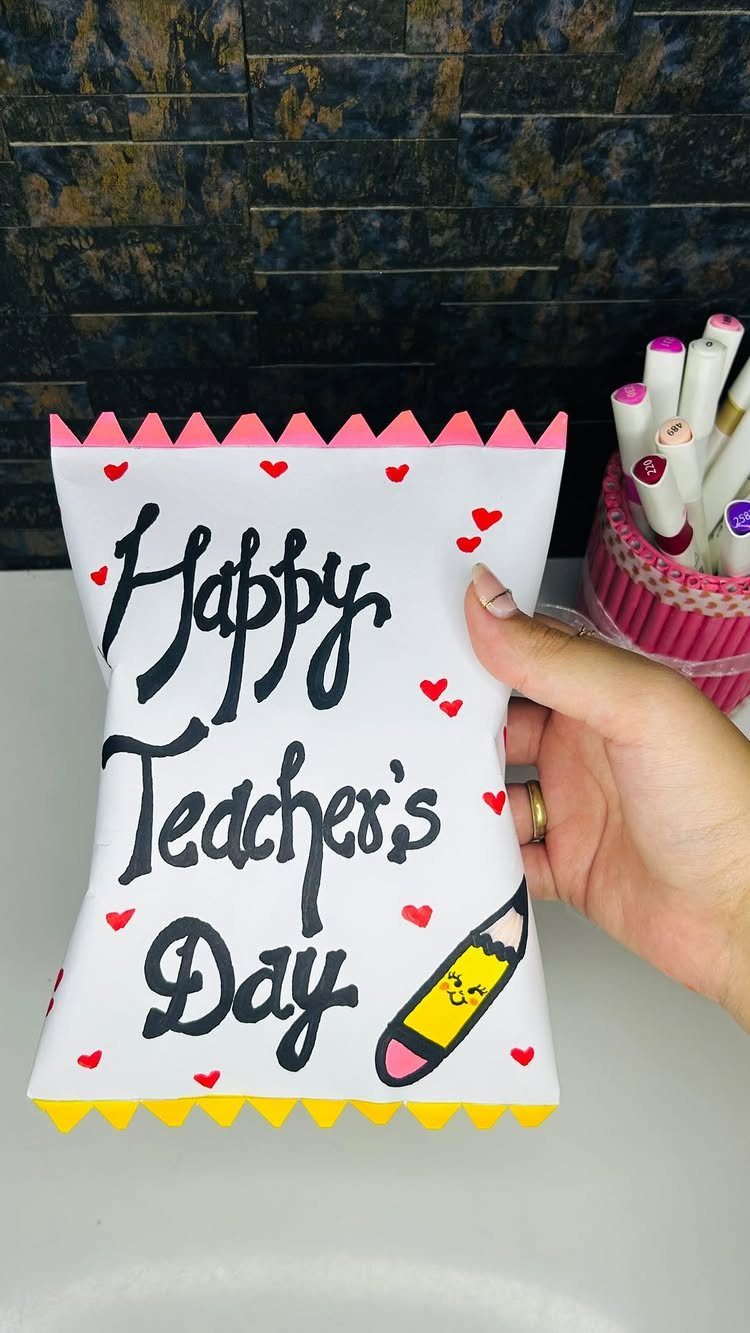 Hand holding a white gift bag with Happy Teacher's Day text and pencil drawing.