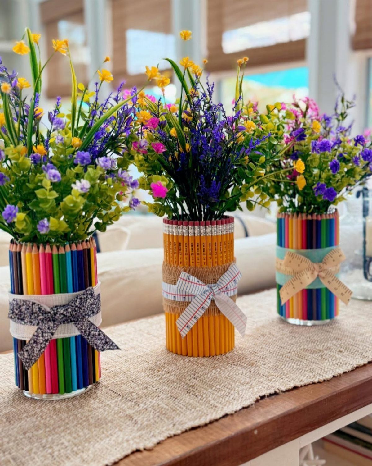 Three vases wrapped in colorful pencils holding bright wildflowers on a burlap runner.