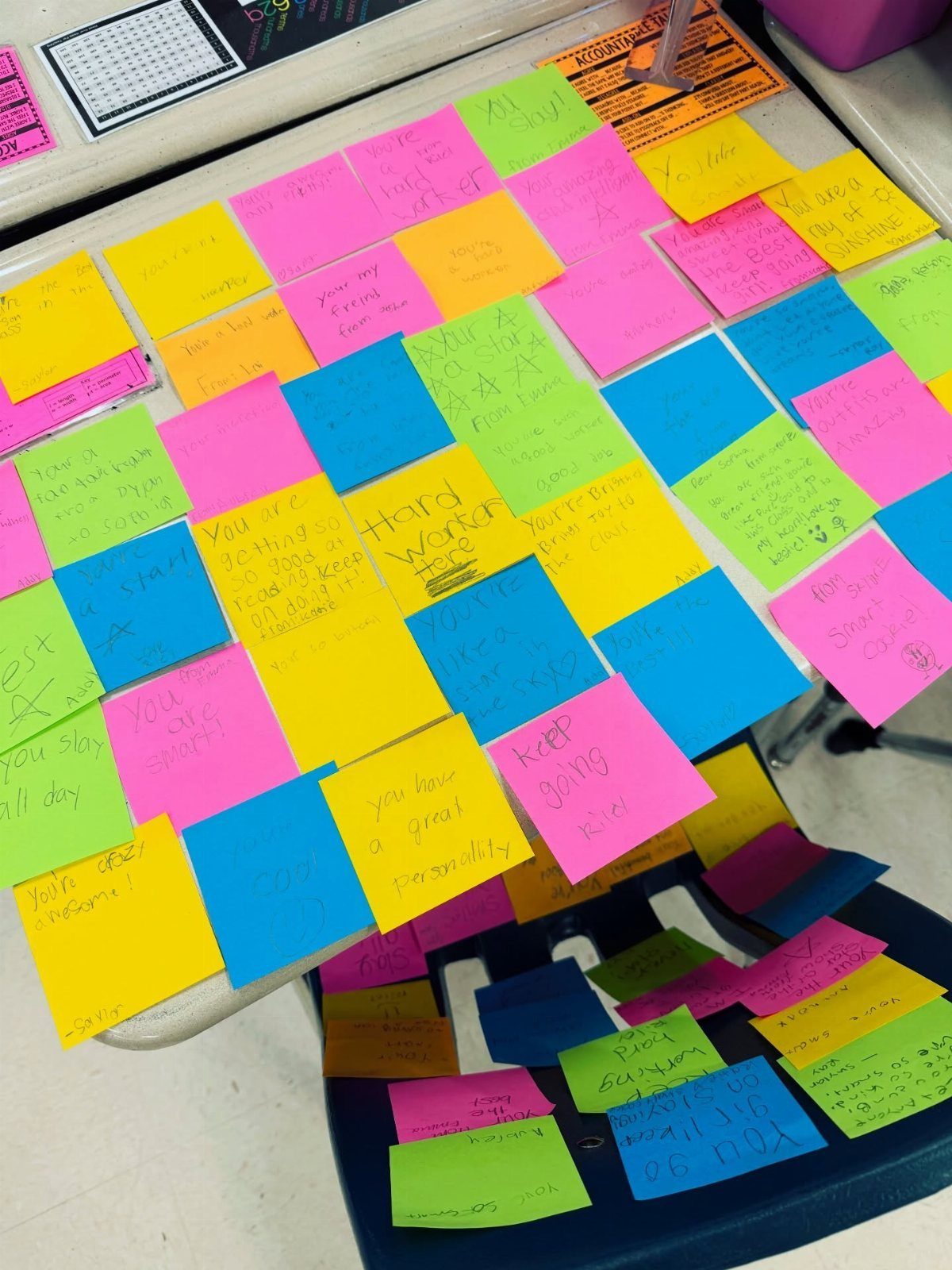 Colorful sticky notes with handwritten compliments arranged on a desk and chair.
