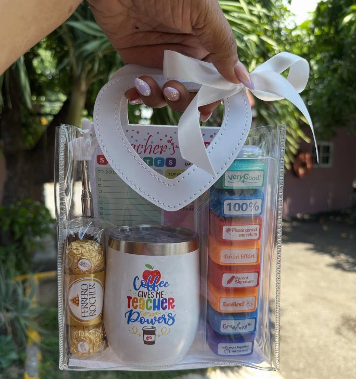 Clear gift bag containing a teacher-themed tumbler, chocolates, stamps, and a notepad.