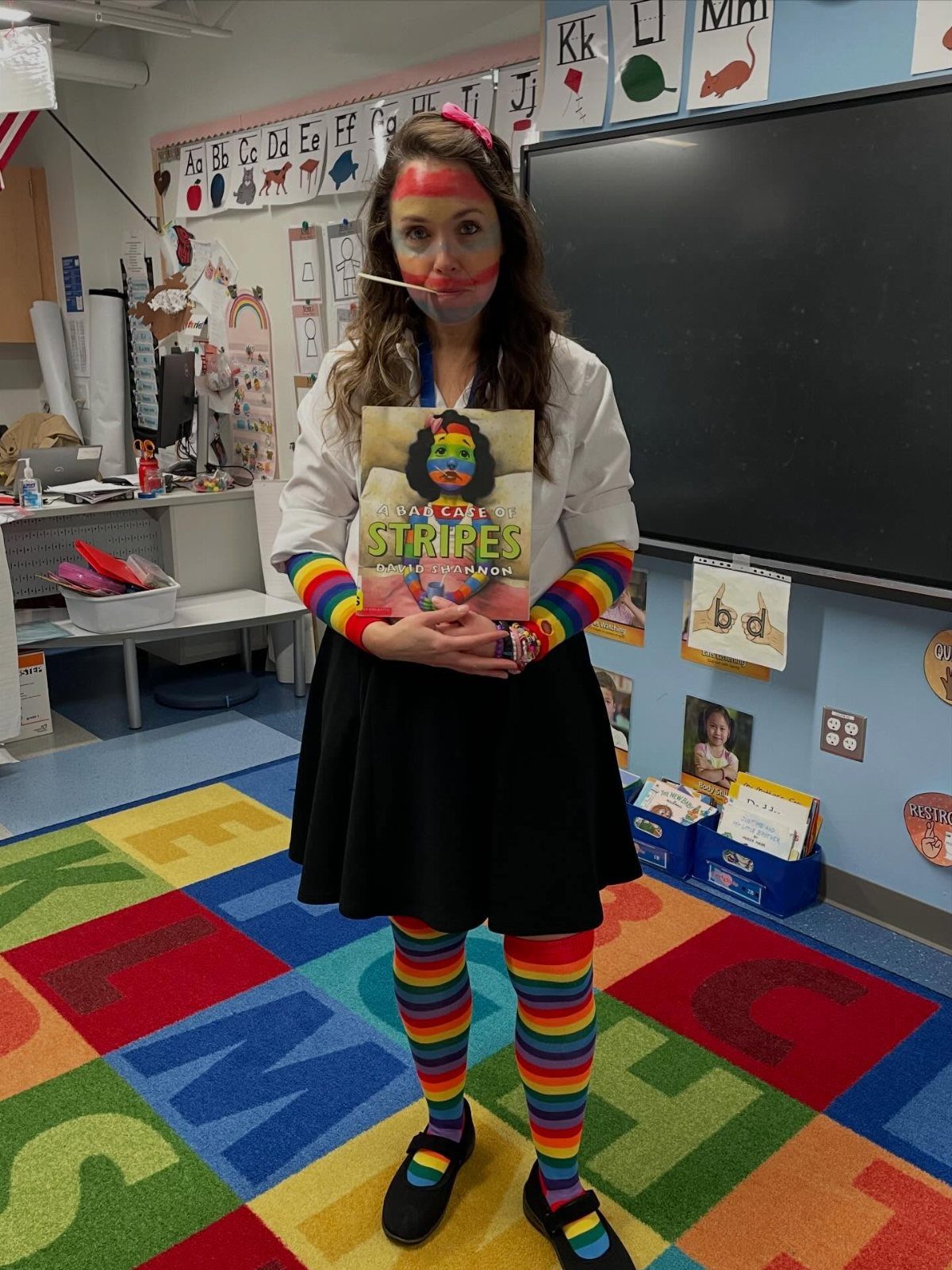 Woman in rainbow tights and face paint holding a children's book in a classroom