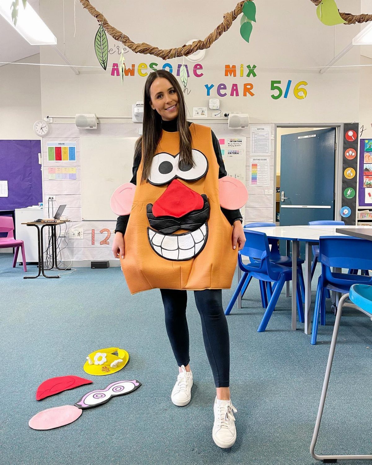 Woman in a classroom wearing a large orange character costume with a smiling face.