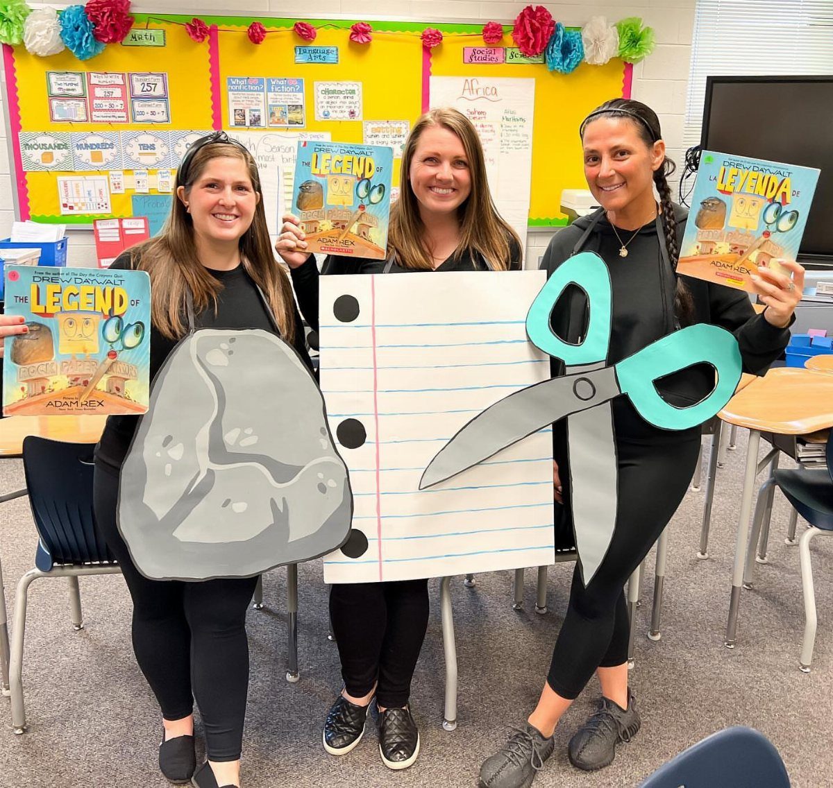 Three women in a classroom wearing rock, paper, and scissors costume cutouts.