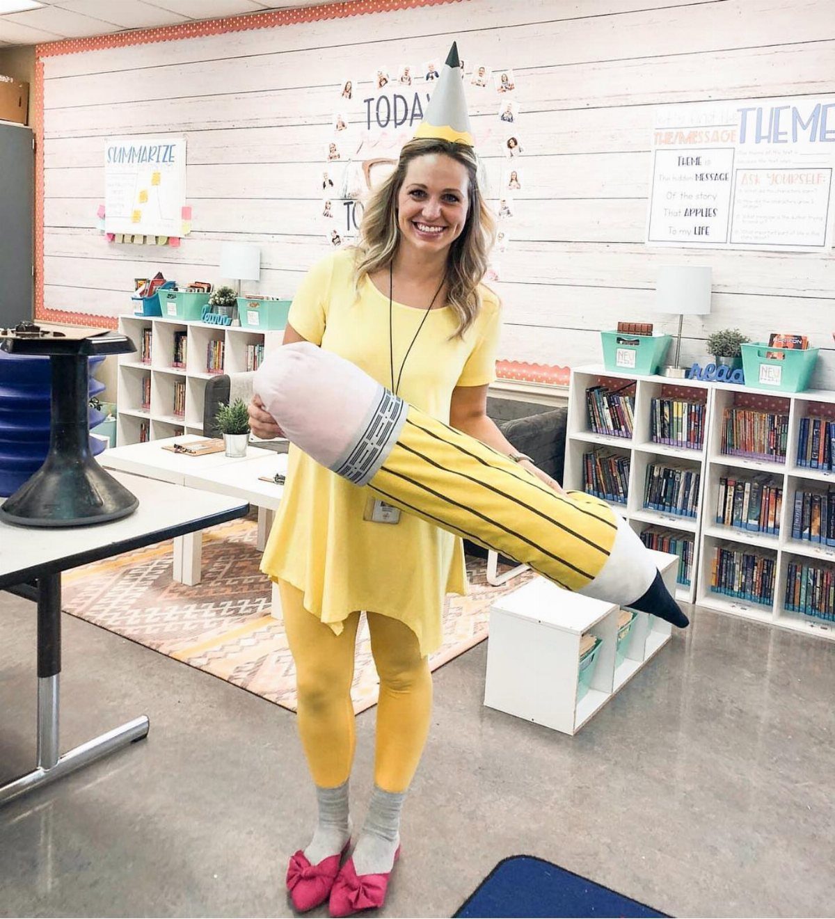 Woman in yellow outfit holding a giant plush pencil in a classroom setting.