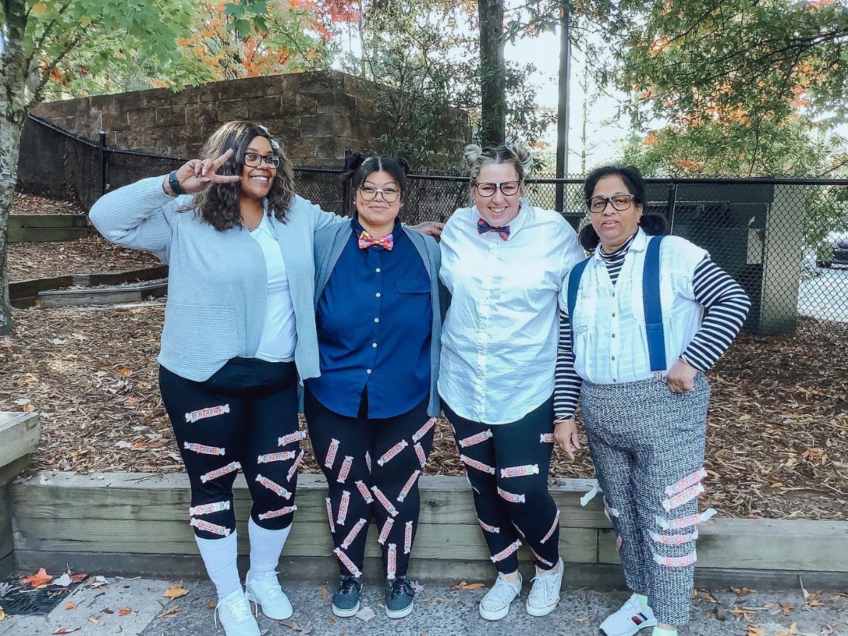 Four women wearing glasses and pants covered in candy wrappers for a costume.