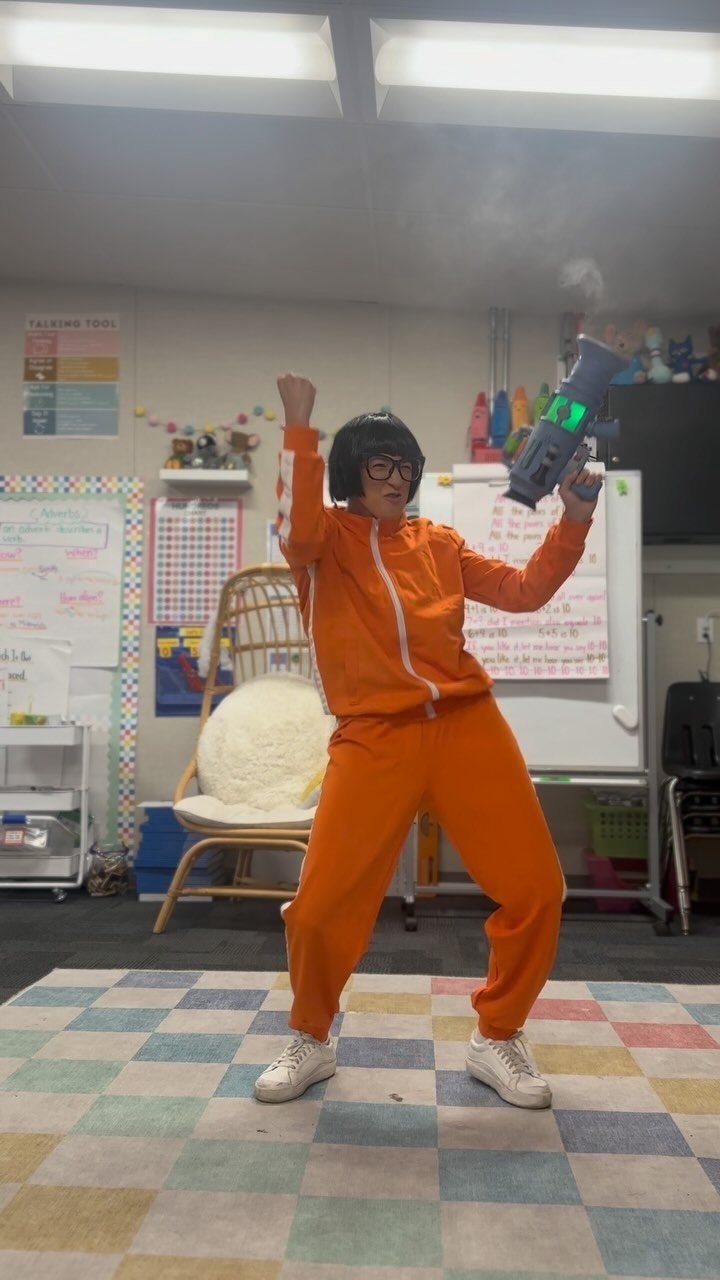 Person in orange tracksuit and black wig holding a smoking prop in classroom
