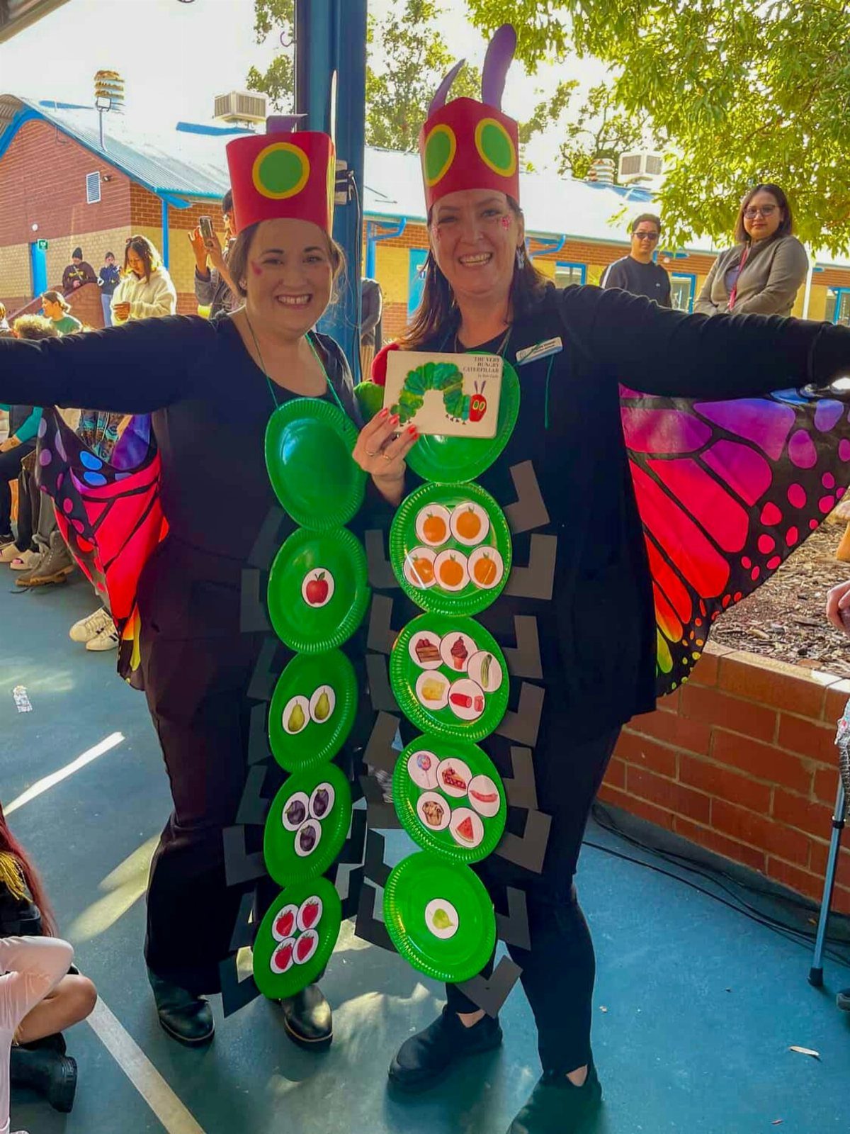 Two women in caterpillar costumes with green plates and colorful butterfly wings.