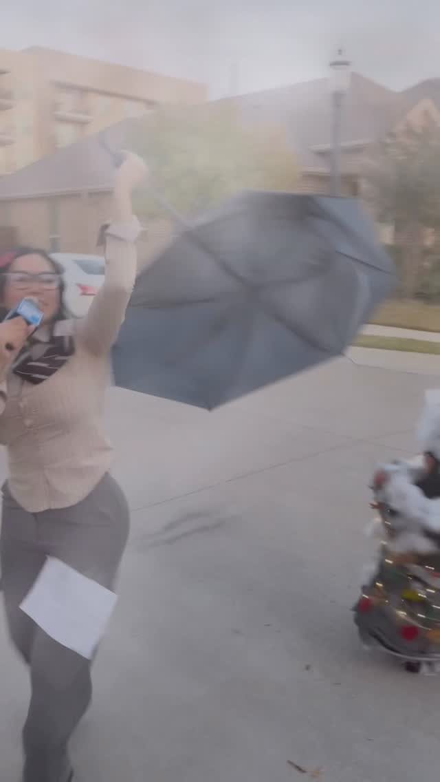 Woman in professional attire holding a microphone and an inside-out black umbrella outdoors.