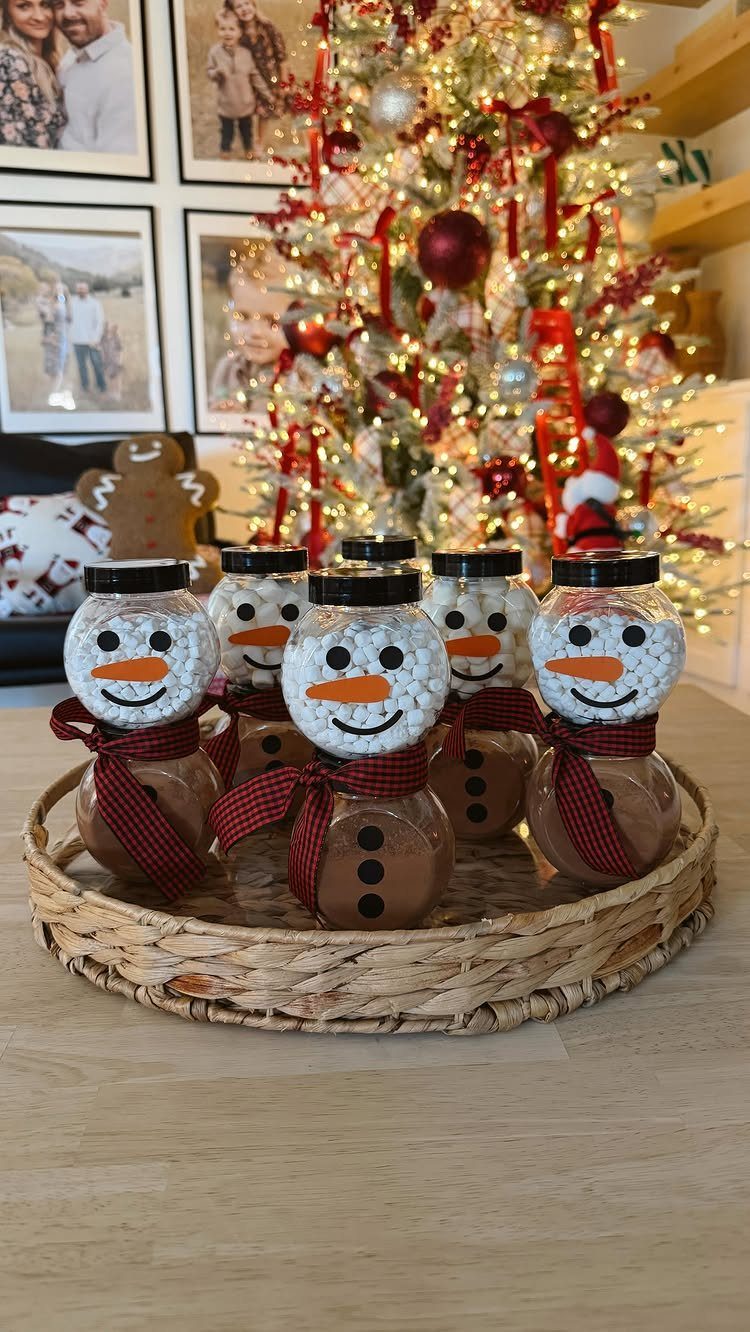 Snowman-themed cocoa jars in a wicker tray before a lit Christmas tree
