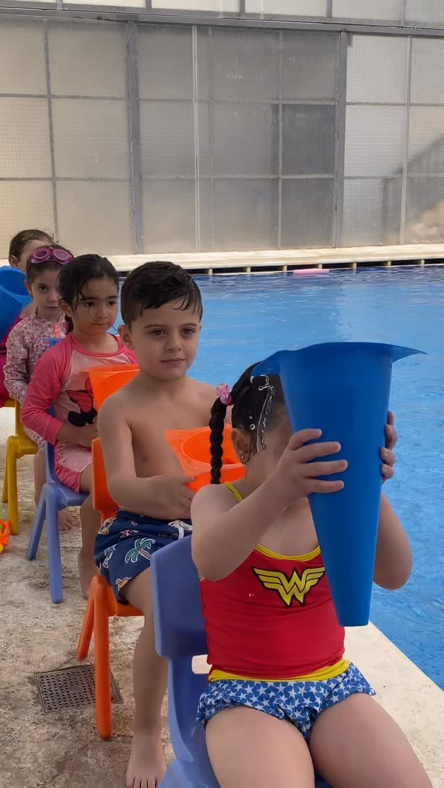Children sitting by a pool holding large colorful plastic funnels for water play.