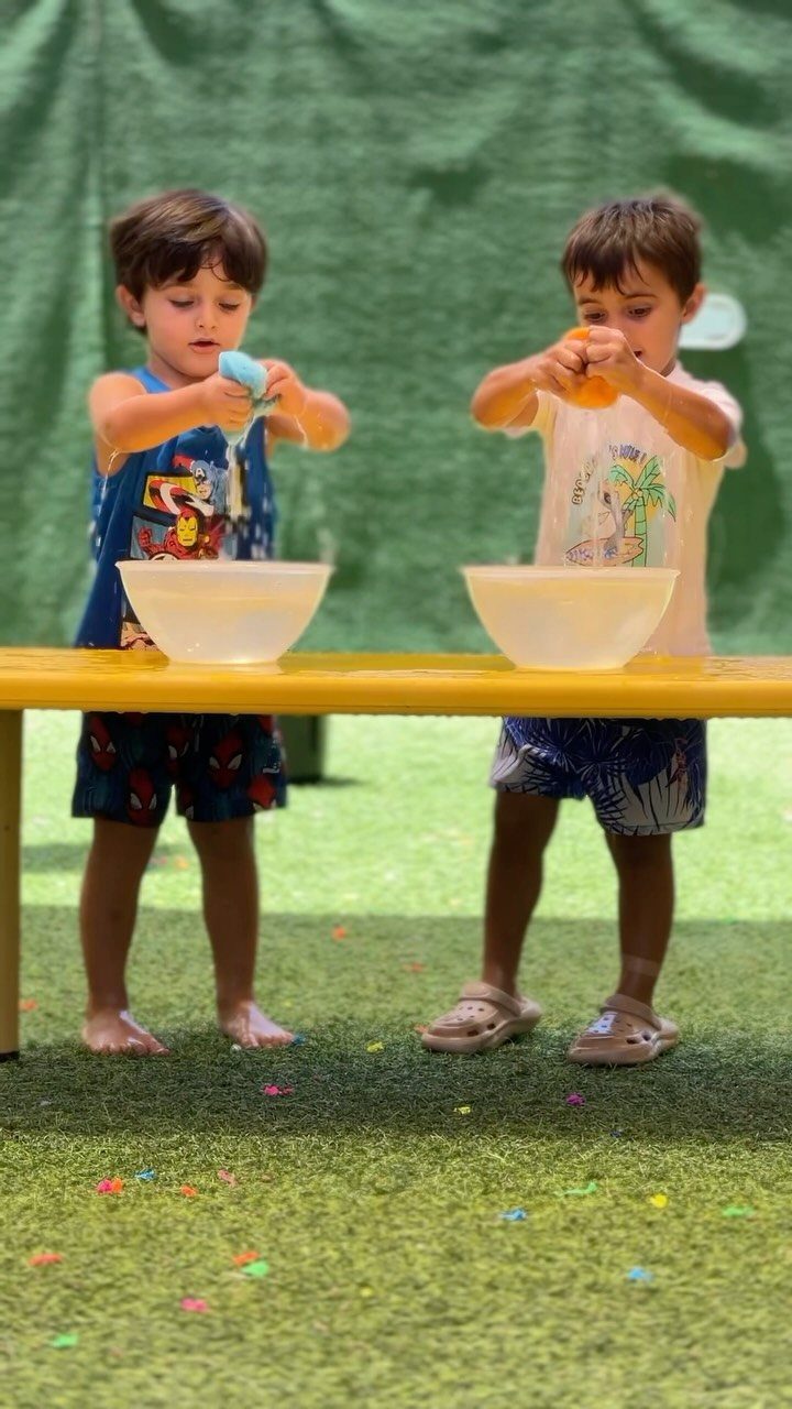Two young boys squeezing colorful sponges into white bowls at a yellow table.