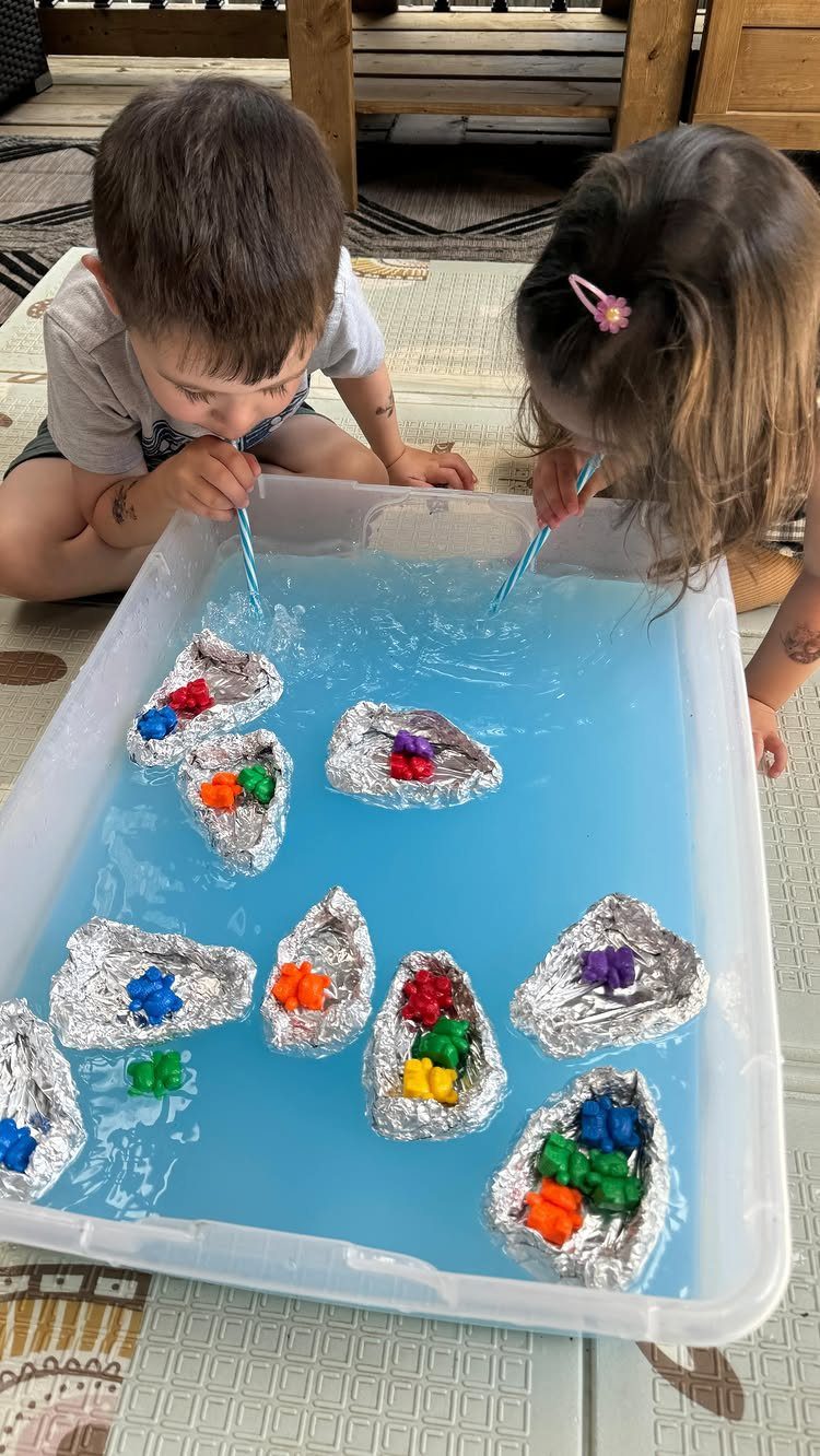 Two children blowing through straws to move foil boats in blue water