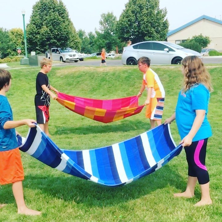 Children holding colorful beach towels taut on a green lawn during outdoor play.