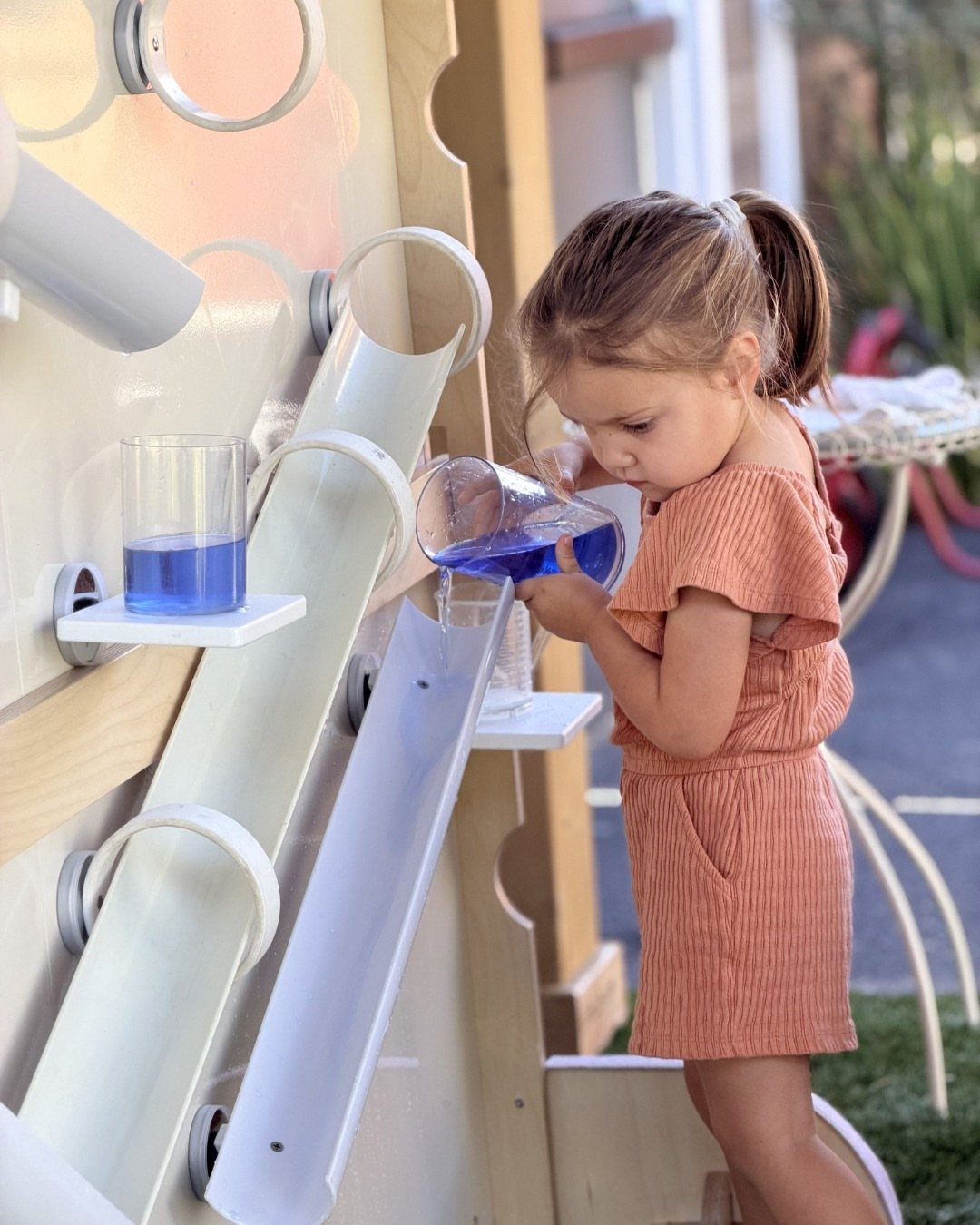 Young girl pouring blue water into a white plastic outdoor water wall activity center.