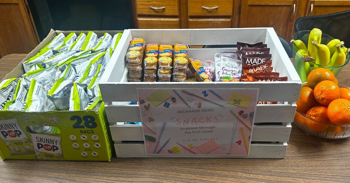 White wooden crate filled with assorted packaged snacks and fresh fruit on table.
